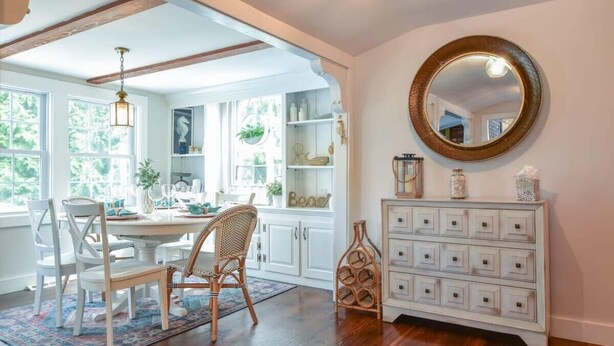 Un coin repas avec une table ronde, des chaises blanches et un plancher en bois. On y trouve un meuble blanc avec des tiroirs et un miroir accroché au mur.
