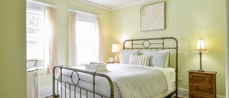 Un dormitorio con estructura metálica para la cama, una mesita de noche con lámpara y una ventana con cortinas.