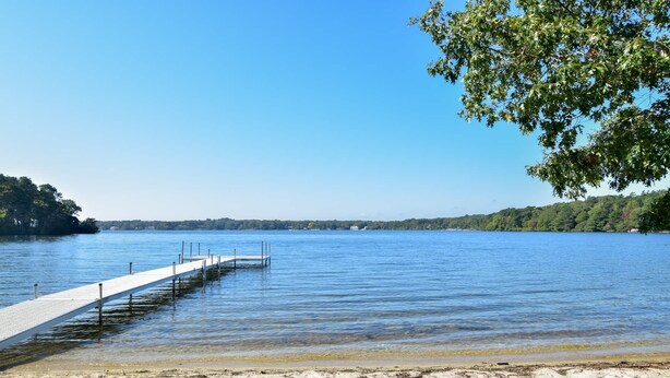 A clear lake with a wooden dock extending into the water, surrounded by trees and a distant shoreline. - Lake House in Centerville with Private Water Access! (Barnstable)