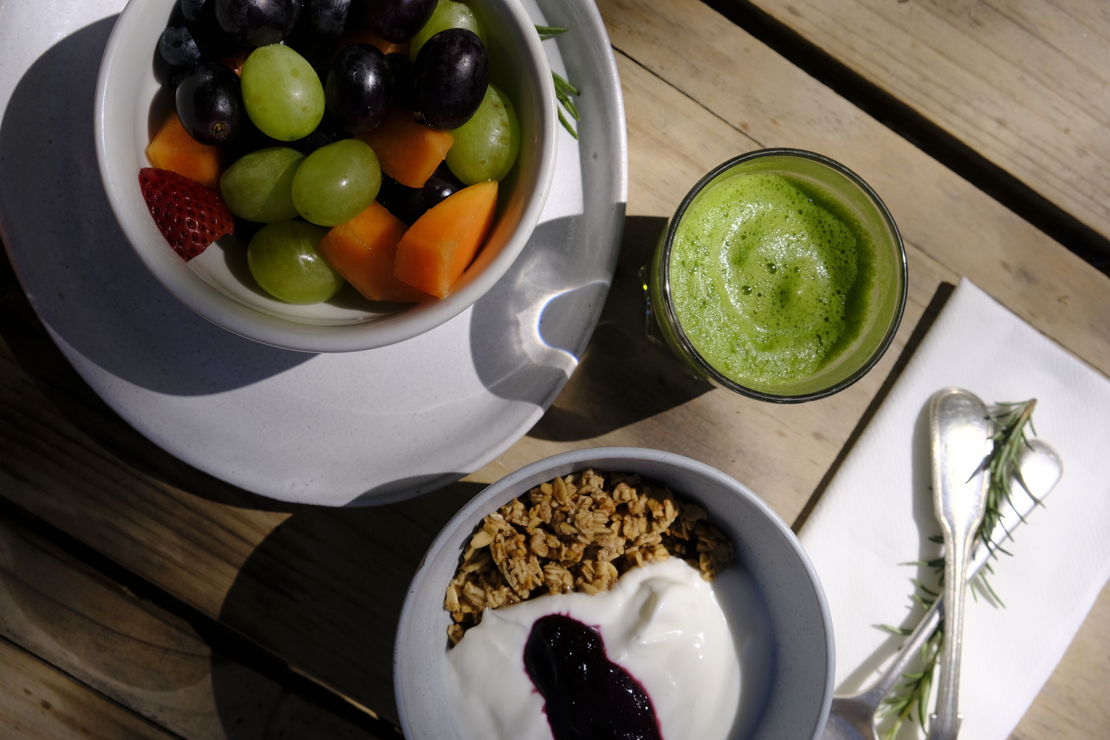 Un repas composé d’un bol de fruits coupés en morceaux, d’un verre de smoothie vert et d’un bol de yaourt agrémenté de granola.