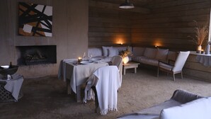 Um interior acolhedor e rústico com lareira, um espaço de jantar montado para dois e uma área de estar confortável com almofadas e um vaso de plantas secas.