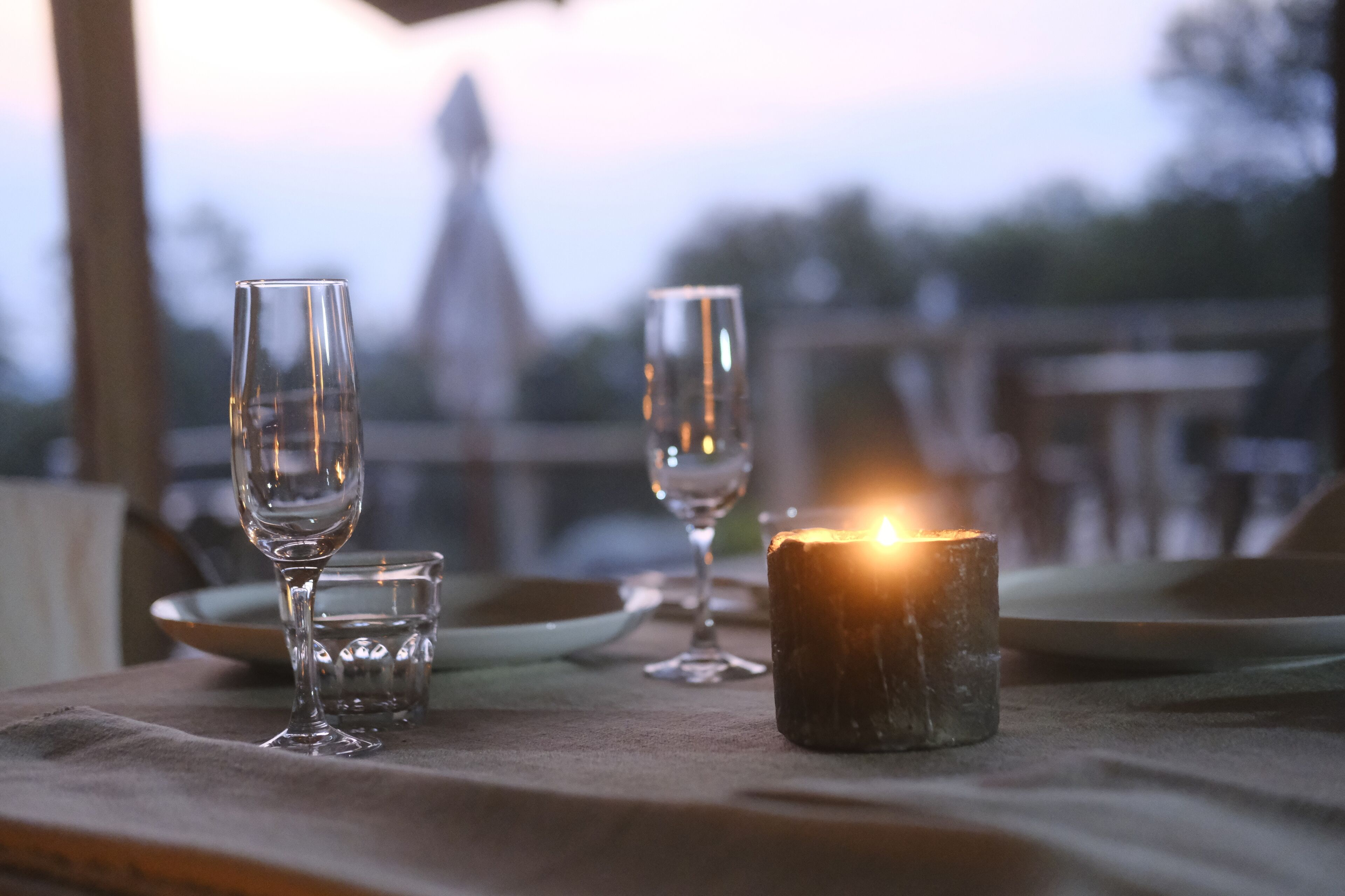 Une table avec trois verres à vin et une bougie allumée, le tout placé sur un fond flou où l’on aperçoit un bâtiment.