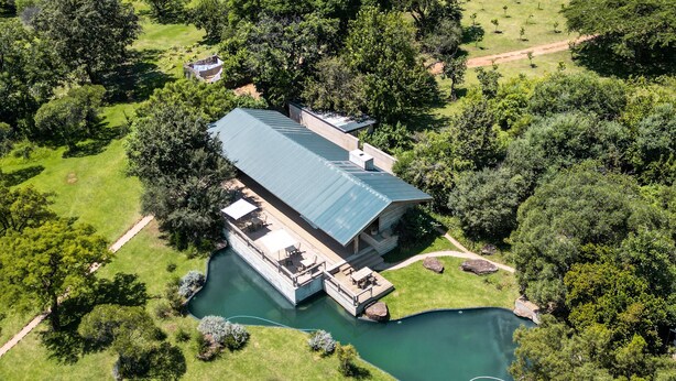 Um edifício grande e moderno com telhado plano, rodeado por vegetação exuberante e um pequeno lago.