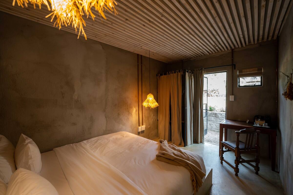 A bedroom with a large bed, a desk, and a chair. There is a window with curtains and a hanging light fixture.