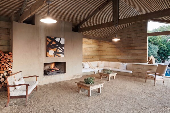 A cozy living area with a fireplace, wooden furniture, and a large window offering a view of greenery. - FARMHOUSE58 (Krugersdorp)