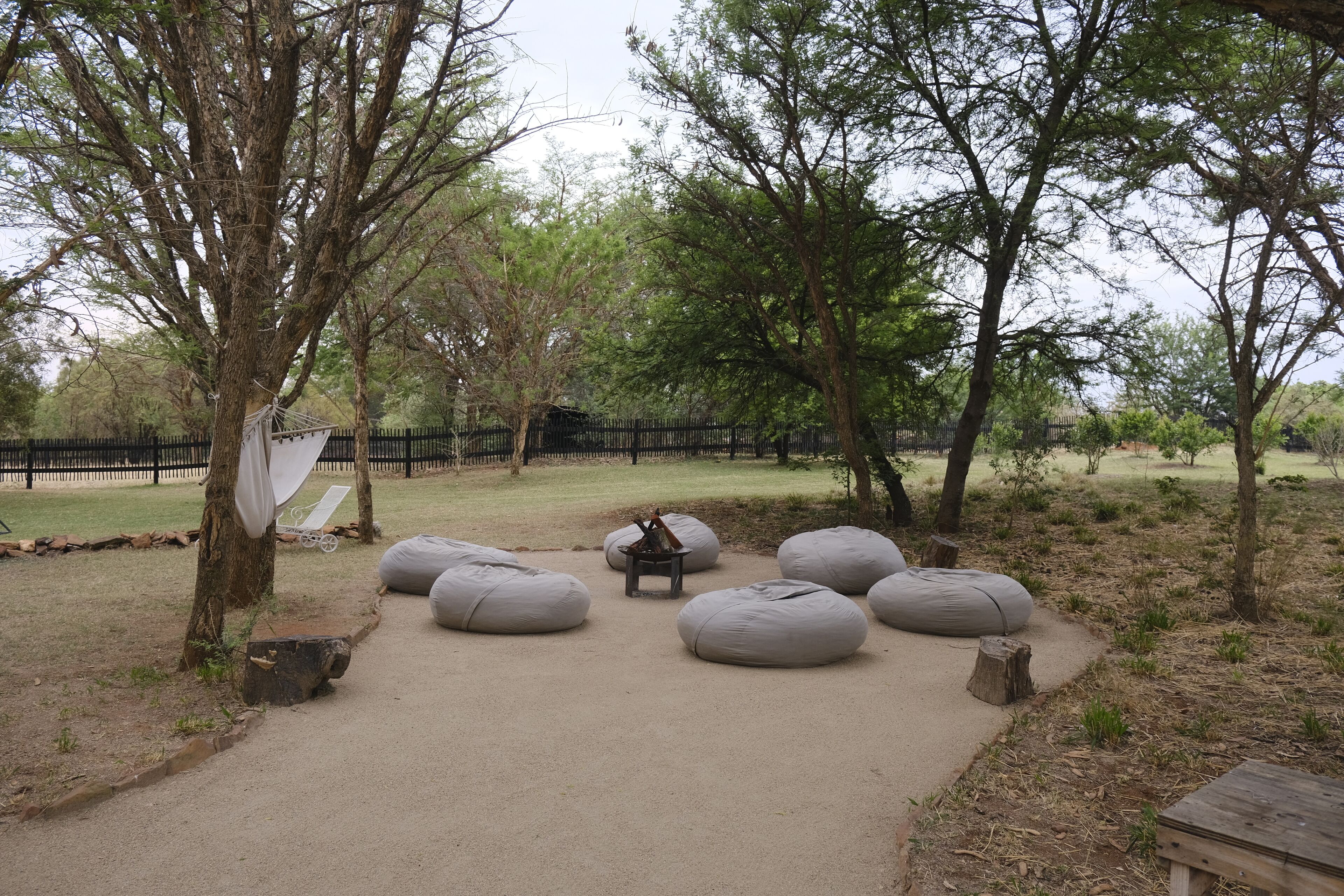 A serene outdoor seating area with large bean bags and a small fire pit, surrounded by trees and a wooden fence.