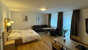 A hotel room with a bed, a sofa, a dining table, and a chair. There is a window with curtains, a plant, and a lamp on the wall. - Motel Zur Dachsbaude (Wandlitz)