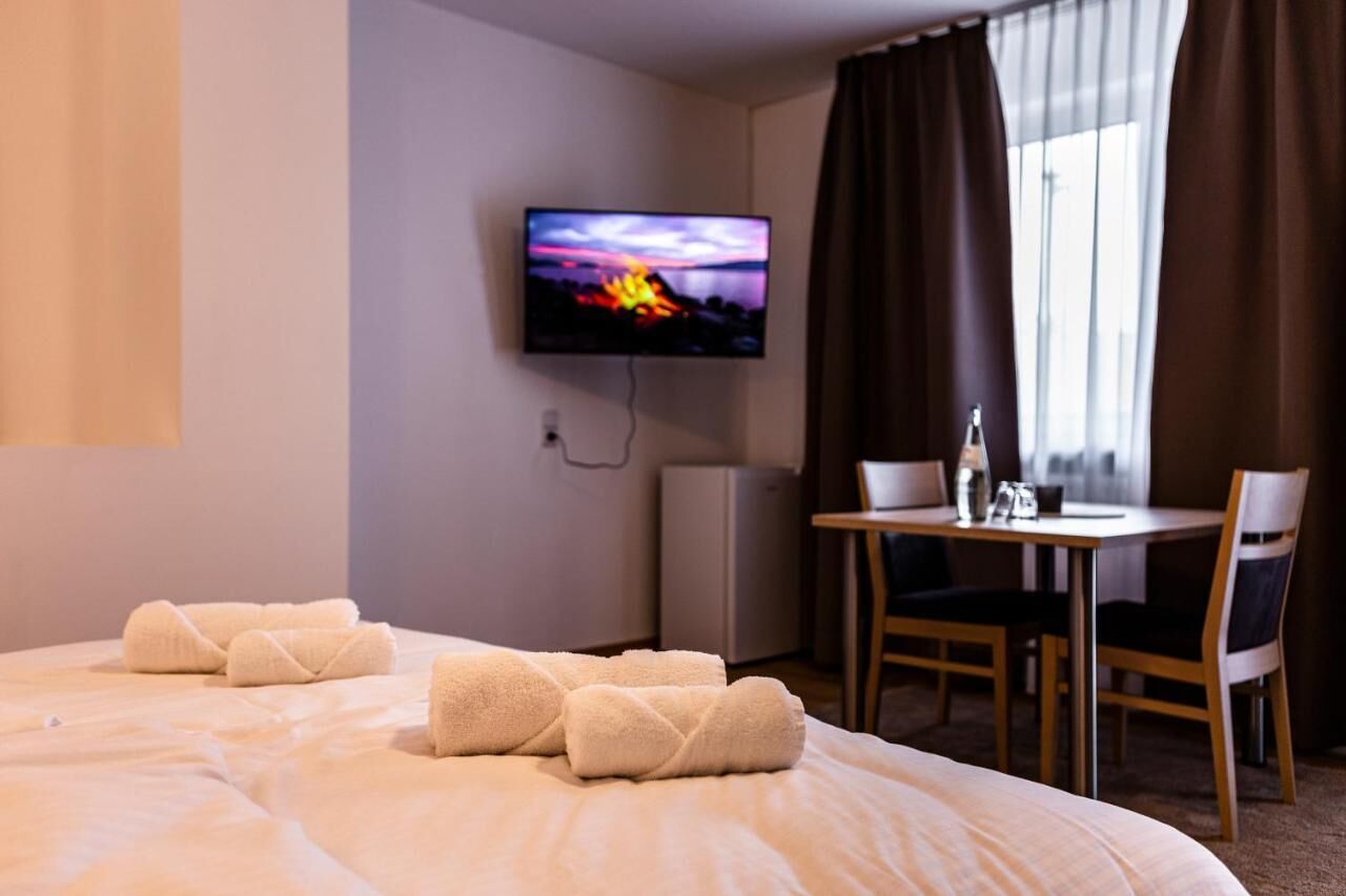 Quarto de hotel com cama, televisão presa na parede, mesinha com cadeiras e janela com cortinas.