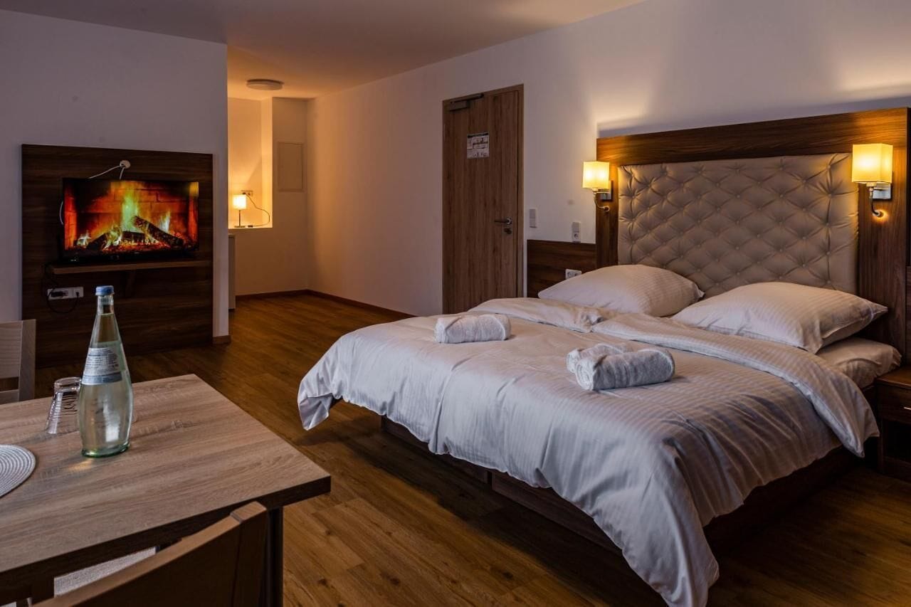 A hotel room with a bed, bedside lamps, a TV on the wall, and a wooden table with a bottle and glasses.