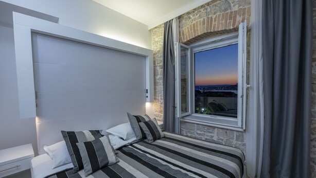 A bedroom with a bed, a window with curtains, a stone wall, and a city view at dusk. - A Heritage Hotel Split (Split)