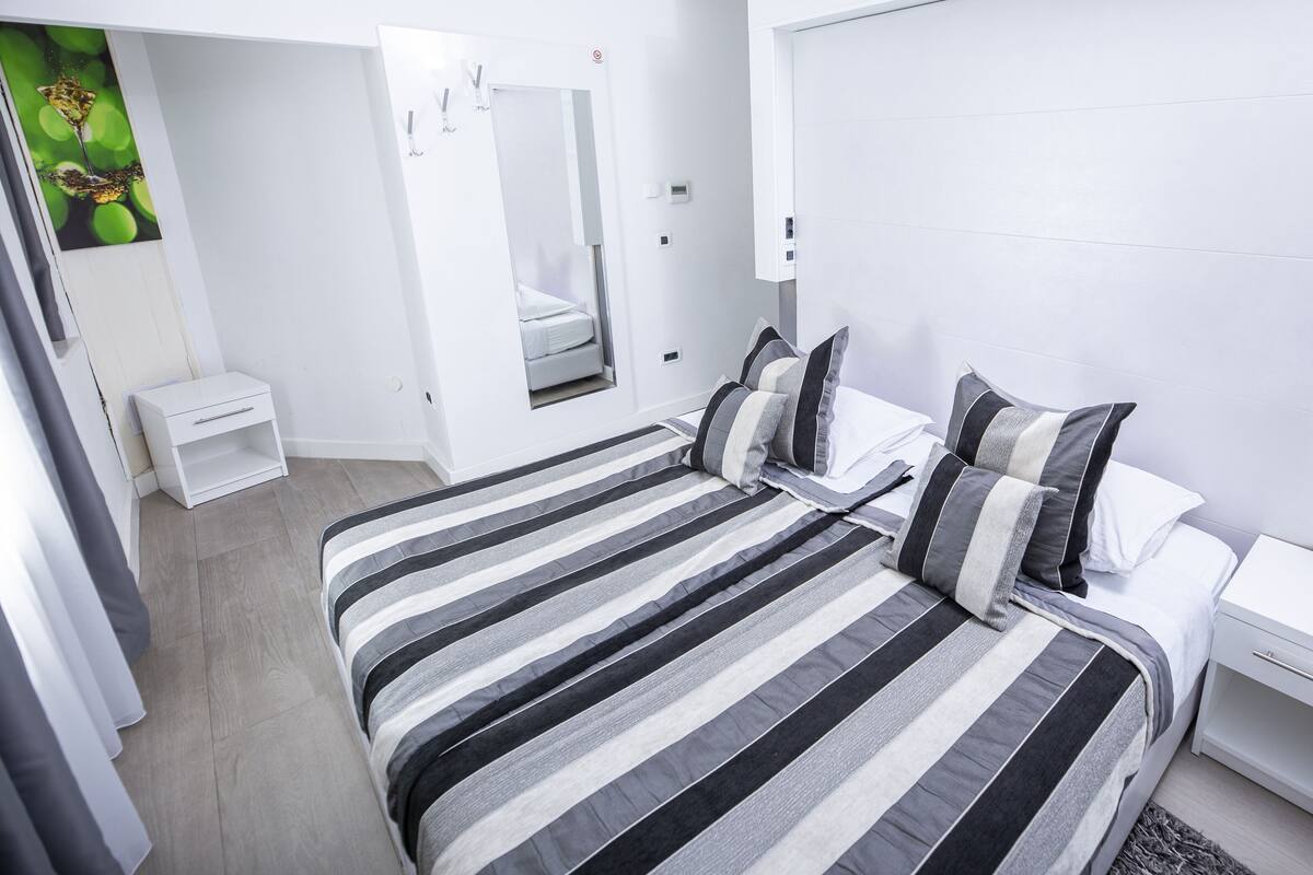 A modern bedroom with a large bed, a bedside table, a wardrobe, and a mirror.