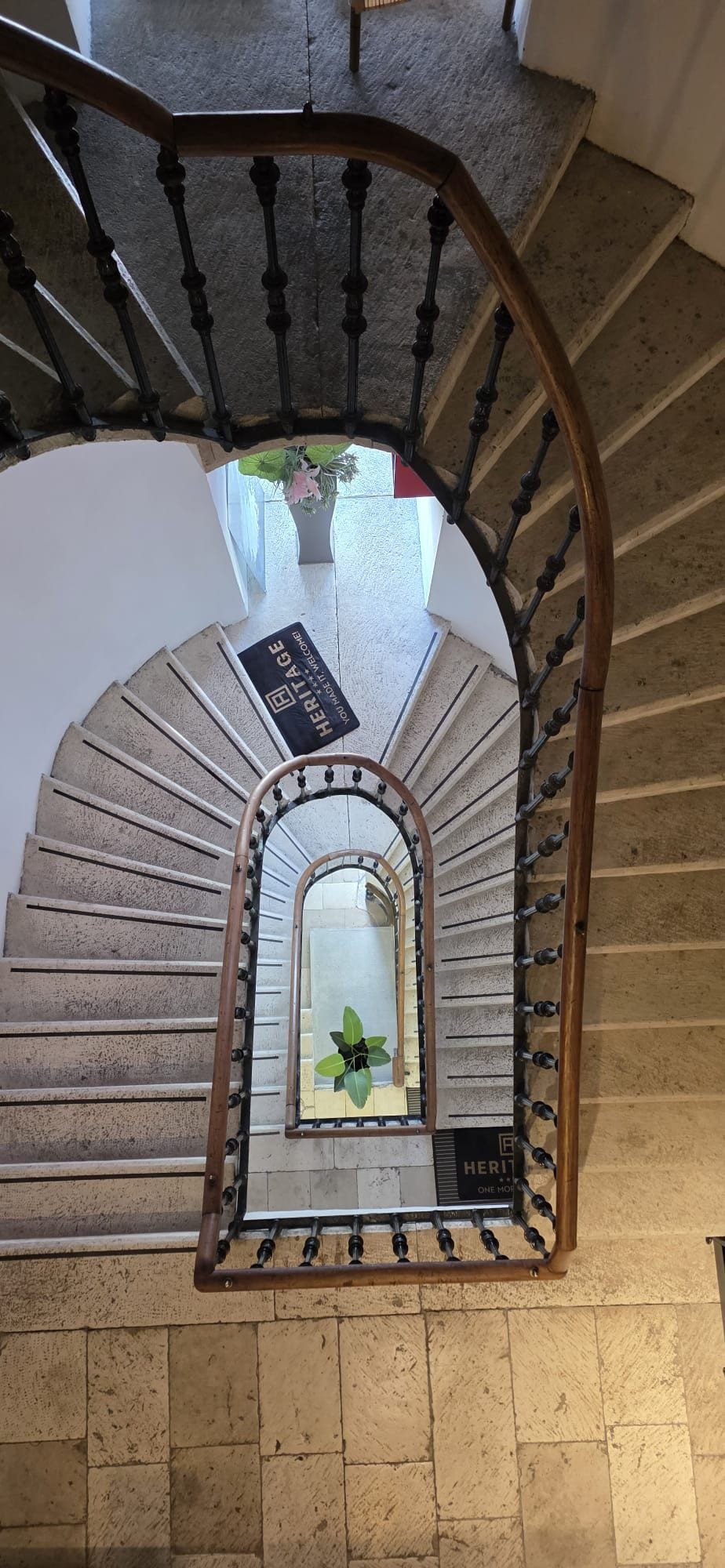 Una scala a chiocciola con ringhiera in legno e balaustre nere, che conduce a un pianerottolo con un cartello che riporta la scritta 'HERITAGE ONE MORE'.