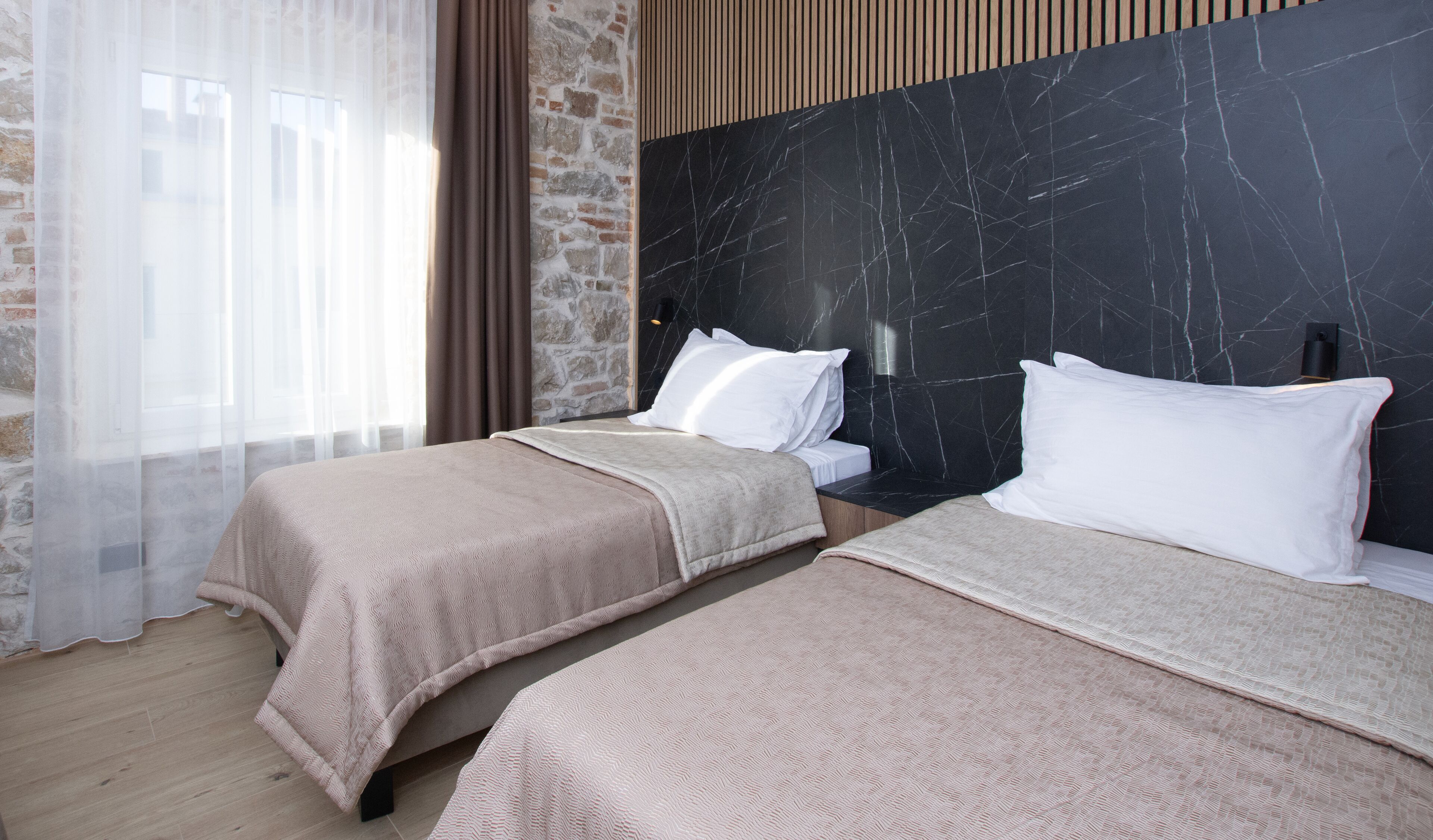 A hotel room with two beds, a stone wall, and a large window with sheer curtains.