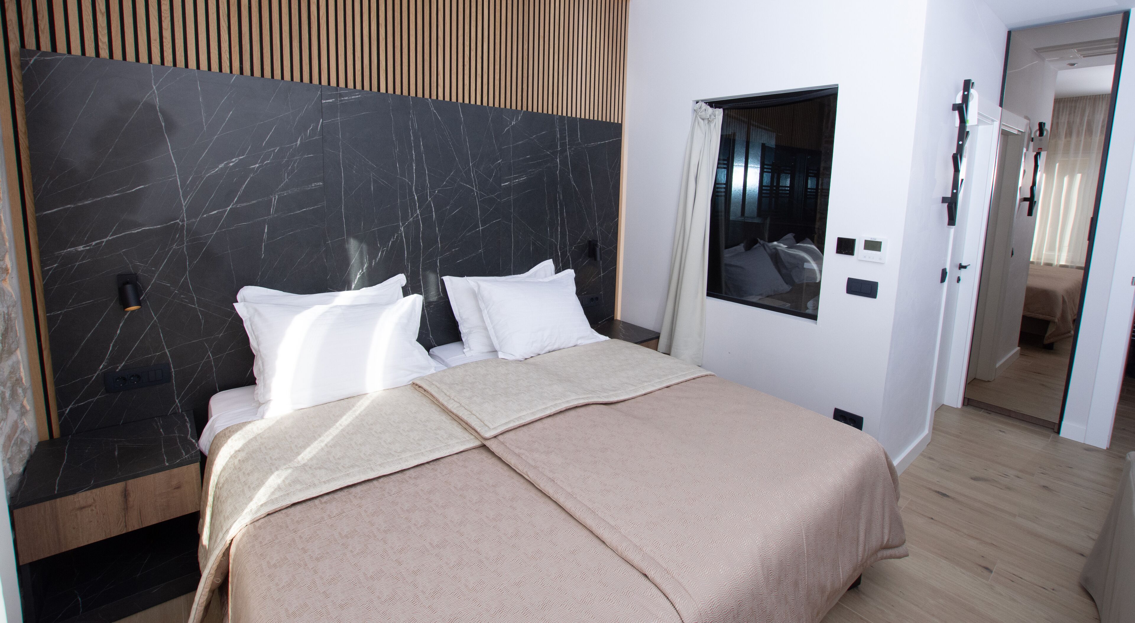 Una habitación de hotel moderna con cabecera de mármol, una cama grande con ropa blanca, una manta color beige y una ventana pequeña que da a la habitación.