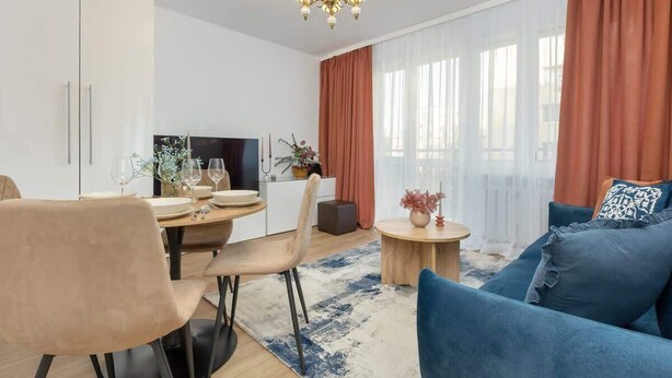 A modern living room with a blue sofa, a dining area with a table set for two, and a kitchen area with a TV and a plant. - Apartment in the center of Warsaw | Balcony and Workspace (Warszawa)