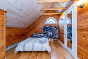 Un dormitorio cálido en el ático, con paredes de madera, techo inclinado, una cama con manta a rayas y dos lámparas fijadas a la pared.