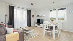 Een moderne woonkamer met een bank, een salontafel, een eettafel met stoelen, een televisie en een groot raam met gordijnen.