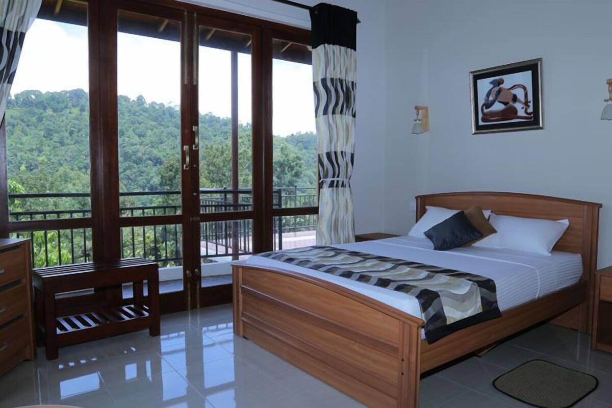 Quarto com uma cama grande, uma mesa de cabeceira de madeira, uma mesinha de apoio, uma cadeira e vista para uma paisagem verde exuberante através de uma porta de vidro deslizante.