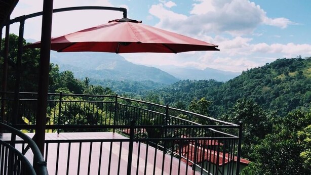 Un balcon agrémenté d’un parasol rouge, d’où l’on peut admirer une vue sur des collines verdoyantes et des montagnes lointaines.