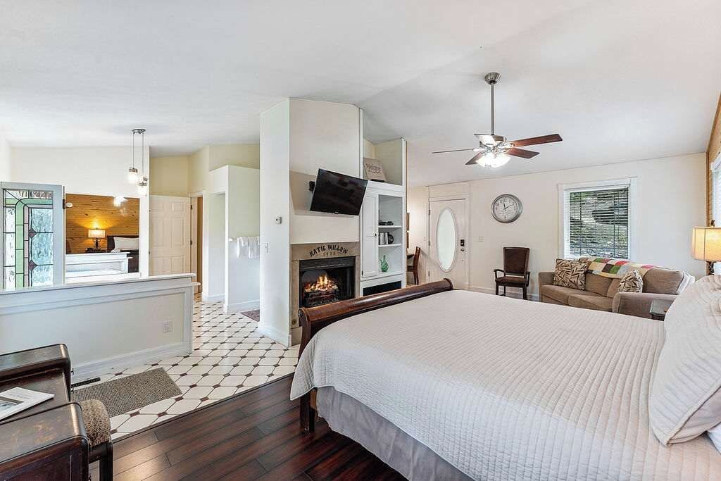 Une chambre avec un grand lit, une cheminée, un ventilateur de plafond et une vue sur le salon.