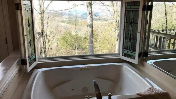 Une salle de bain avec une grande baignoire, une fenêtre donnant sur des arbres et des montagnes, et une terrasse en bois à l’extérieur.