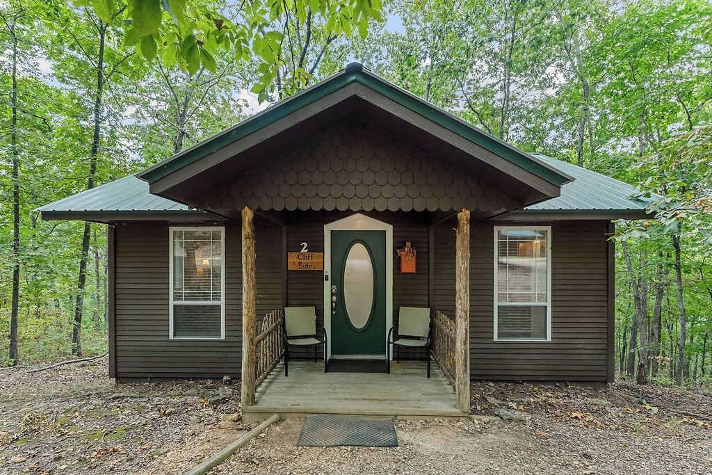 A cabin with a green door, two windows, and a porch with chairs.