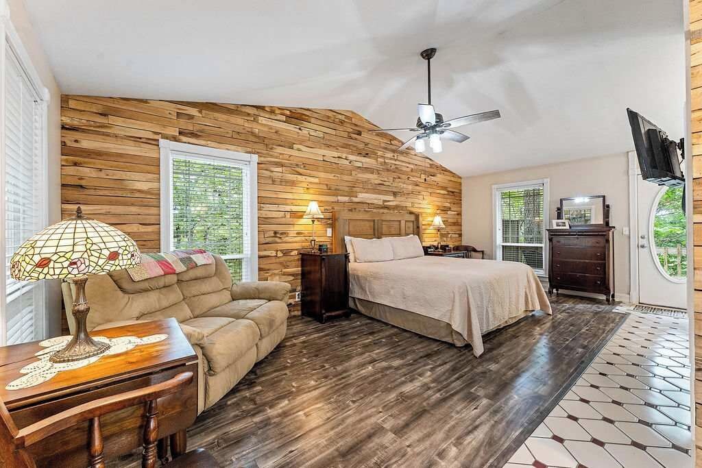 A bedroom with a wooden wall, a bed, a sofa, a nightstand, a lamp, and a ceiling fan.