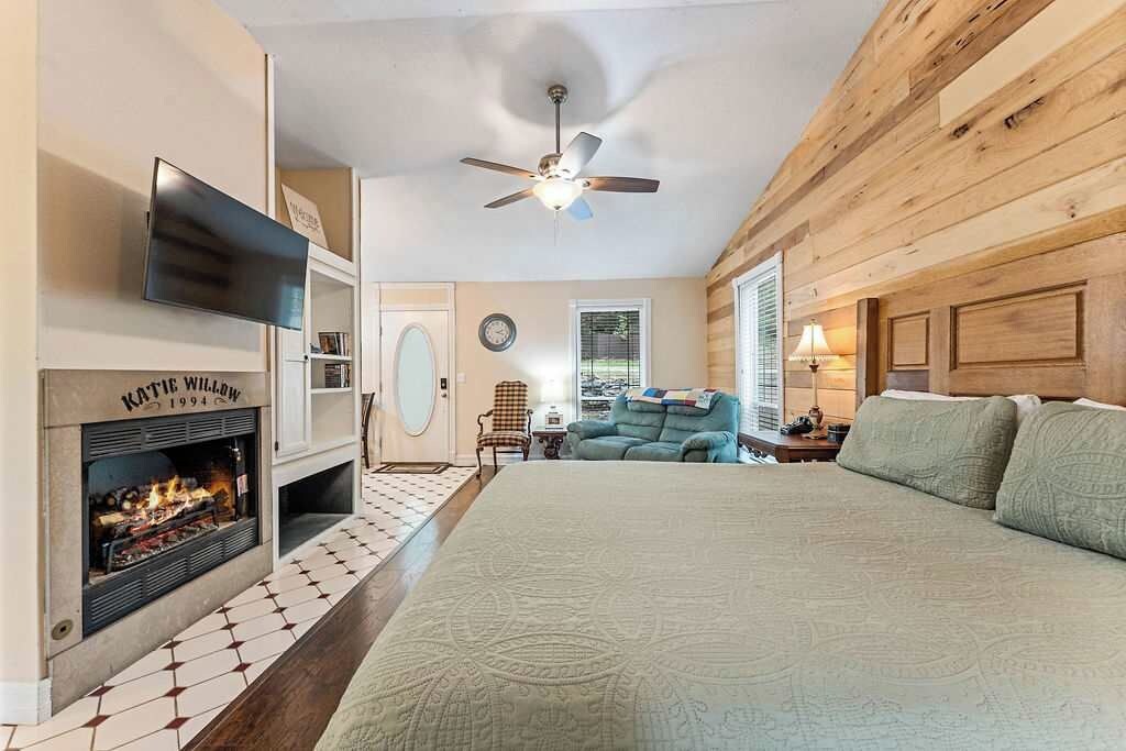 A bedroom with a fireplace, a bed, a ceiling fan, and a view of the outdoors.
