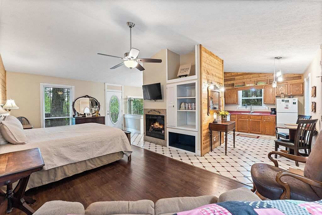 A bedroom with a bed, a desk, and a chair. There is a fireplace, a television, and a kitchen area visible in the background.