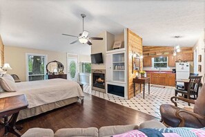 A bedroom with a bed, a desk, and a chair. There is a fireplace, a television, and a kitchen area visible in the background. - Ozark Spring Cabin 04 Firefly Glow, King, Spa Tub (Eureka Springs)