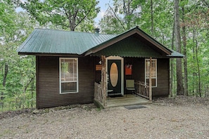 A rustic cabin with a porch, a door with a circular window, and a small set of stairs leading up to the entrance. - Ozark Spring Cabin 04 Firefly Glow, King, Spa Tub (Eureka Springs)