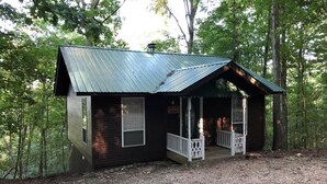 A small cabin with a metal roof, a porch, and a gravel driveway surrounded by trees. - Ozark Spring Cabin 04 Firefly Glow, King, Spa Tub (Eureka Springs)