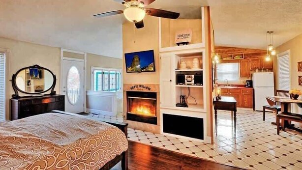 A bedroom with a bed, a ceiling fan, a fireplace, a kitchen area with cabinets and a refrigerator, and a dining area with a table and chairs. - Ozark Spring Cabin 04 Firefly Glow, King, Spa Tub (Eureka Springs)