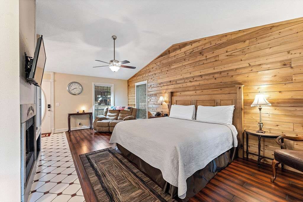 A bedroom with a wooden sloped ceiling, a bed with white bedding, a ceiling fan, a fireplace, and a patterned rug.