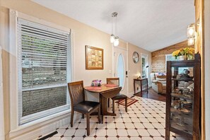A dining area with a table and chairs, a window with blinds, a clock on the wall, and a wooden cabinet with glassware. - Romantic Ozark Cabin | Spa Tub • Trails • Cave (Eureka Springs)