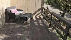 Een houten terras met een rieten stoel en een klein tafeltje, een houten leuning en een boom op de achtergrond.