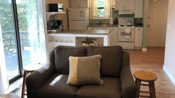 Een moderne woonkamer met een bank en een klein krukje, een keuken met witte kasten en een raam, en een schuifpui die leidt naar een terras.