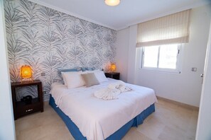 A bedroom with a bed, two bedside tables, a window with blinds, and a wall with a leaf pattern.