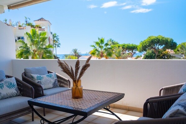 An outdoor seating area with wicker furniture and a table, overlooking a coastal landscape with palm trees and buildings.