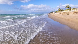 A beach with waves, a sandy shore, and a building with a fence and palm trees.