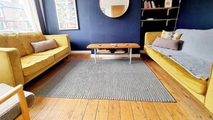 Ein modernes Wohnzimmer mit einem gelben Sofa, einem runden Spiegel, einem Bücherregal und einem gemusterten Teppich.
