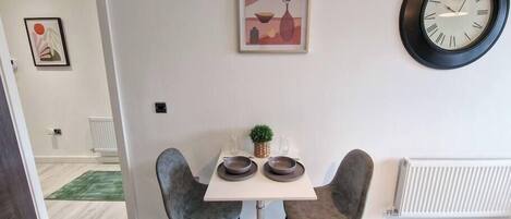 A dining area with a table, two chairs, a clock, and a framed picture on the wall.