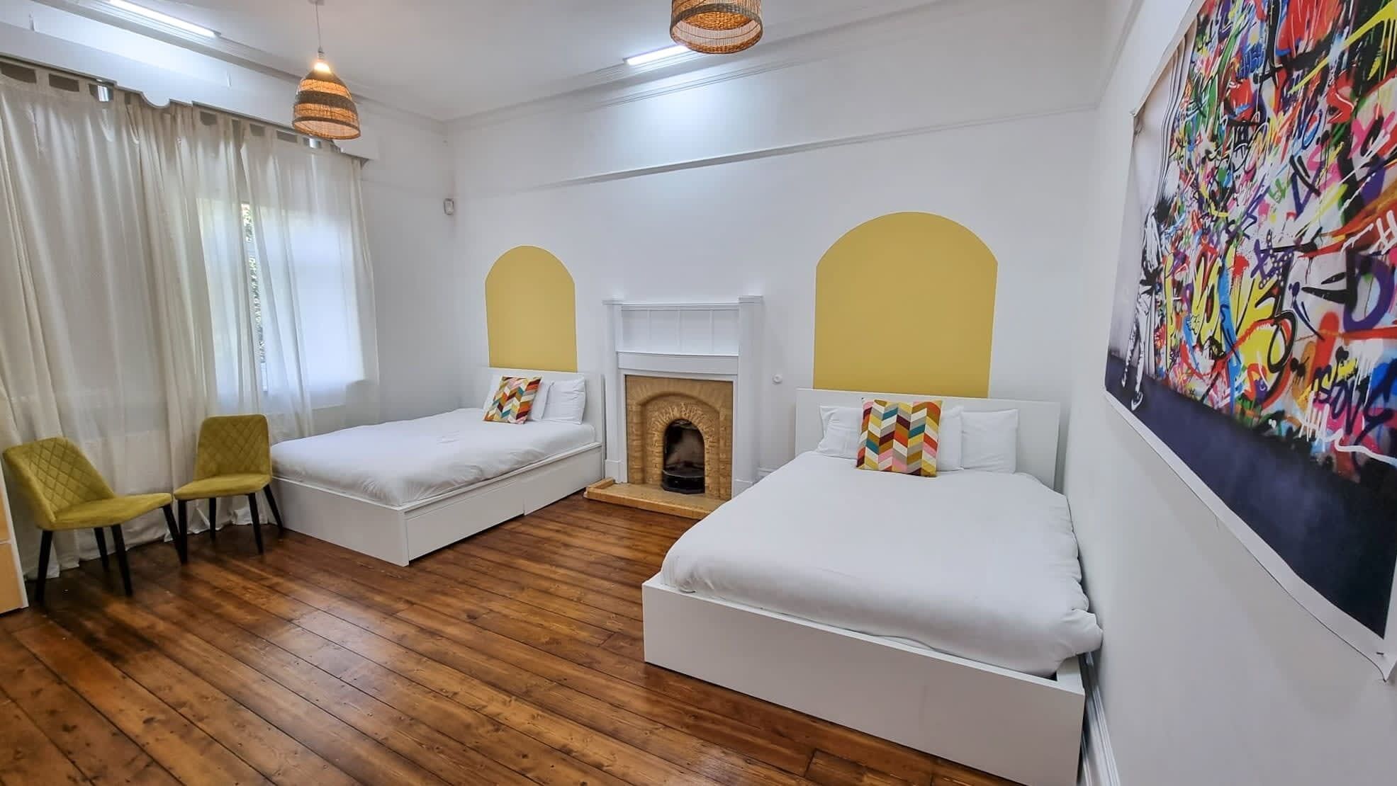 A room with two beds, a fireplace, a yellow chair, and a colorful wall mural.