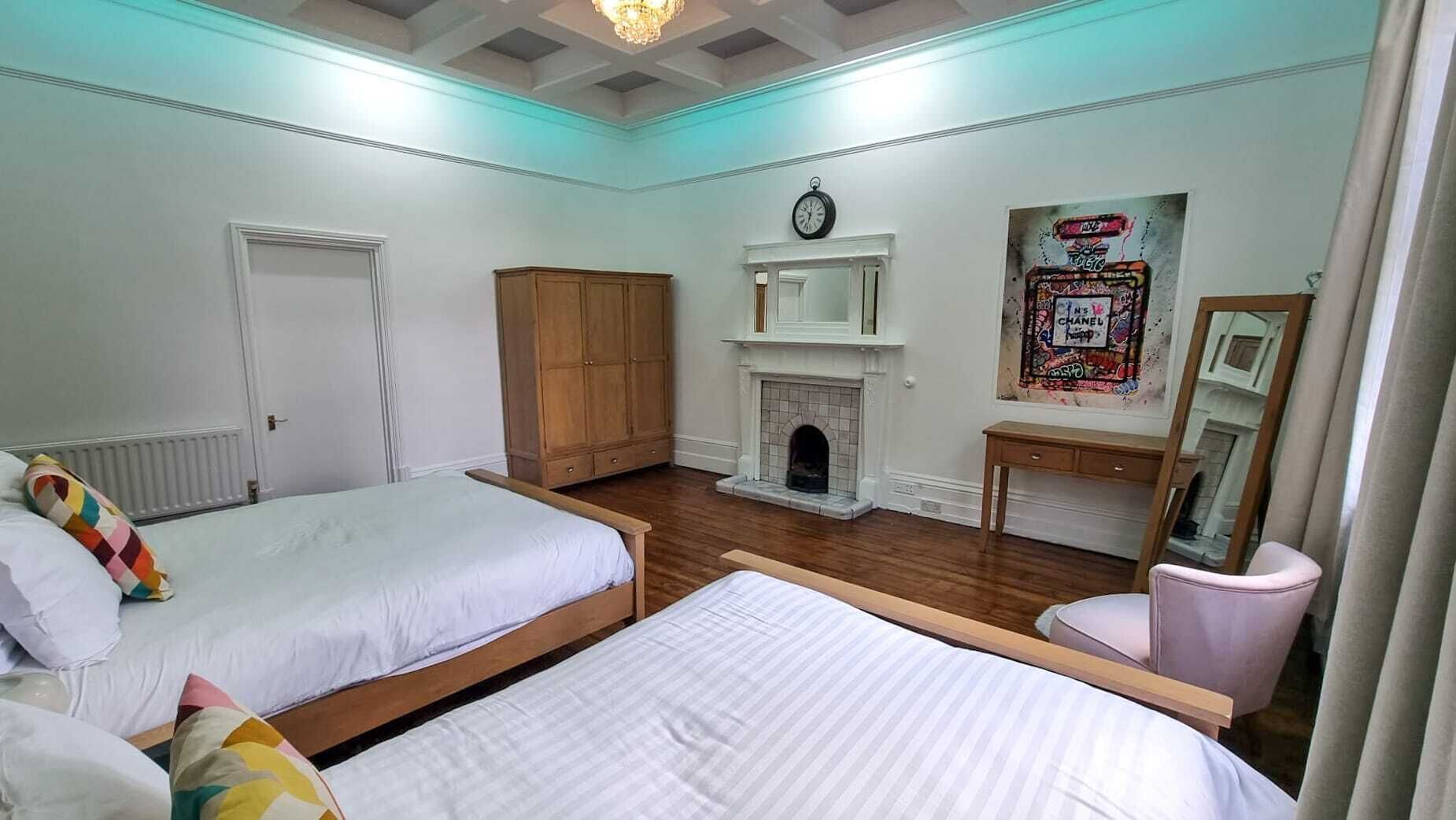 A bedroom with two beds, a wooden wardrobe, a fireplace, a clock, and a mirror.