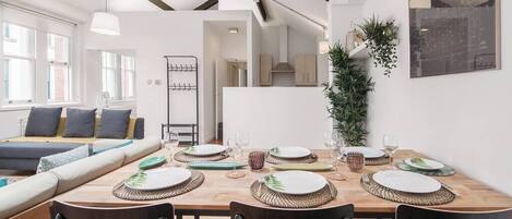 Un comedor moderno con una mesa de madera dispuesta para seis comensales, un sofá al fondo y una cocina visible a través de un vano abierto.