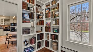 Una biblioteca amplia con estanterías blancas llenas de libros e imágenes enmarcadas, un comedor con una mesa redonda y sillas, y un gran ventanal con vista a árboles desnudos.