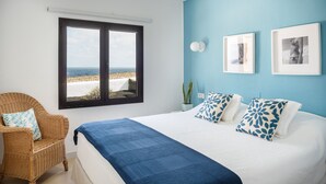 Un dormitorio con una cama grande, una silla de mimbre, un ventanal con vista a la playa y dos cuadros en la pared.