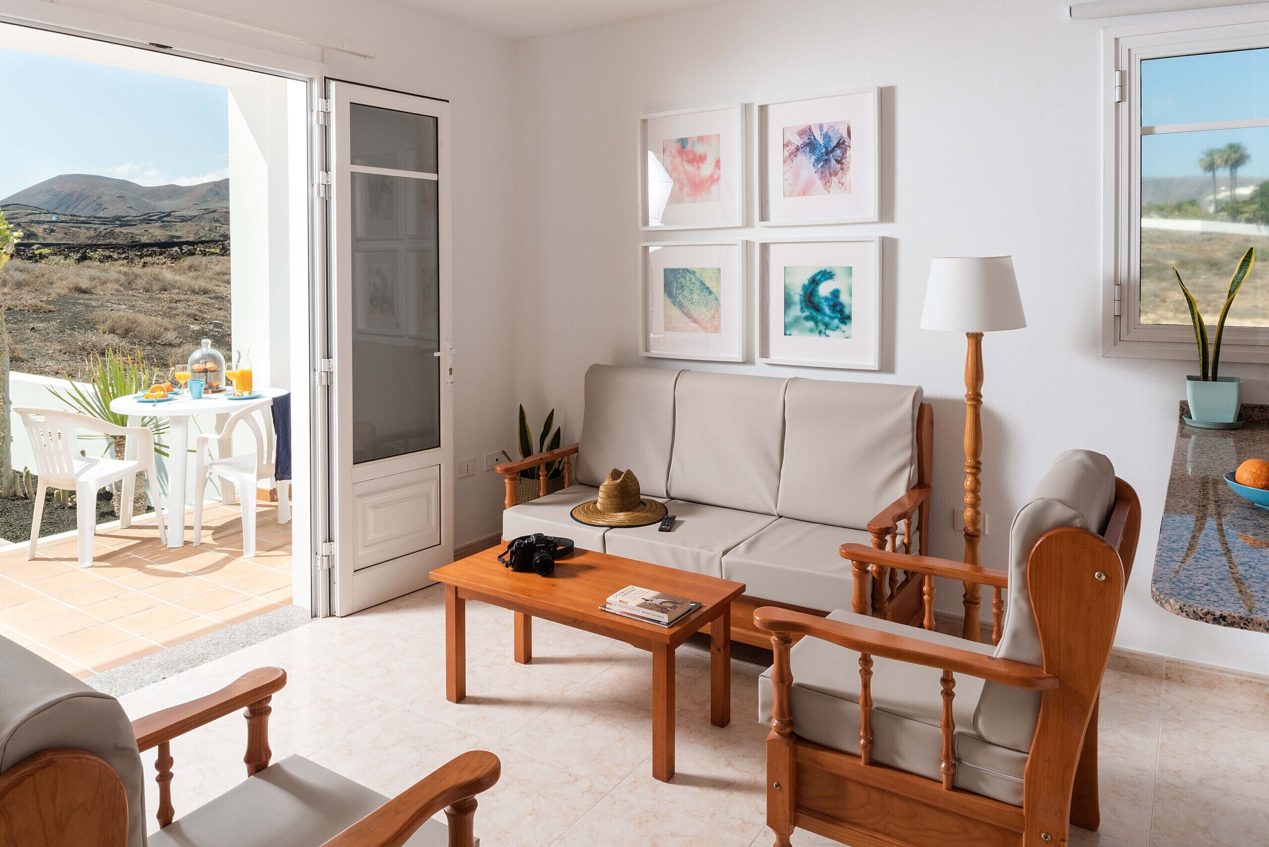 A living room with a sofa, wooden chairs, a coffee table, and a dining table with chairs. There are framed artworks on the wall and a lamp.