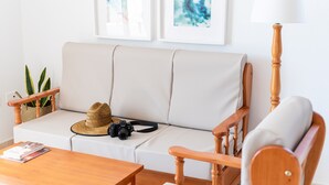 Ein gemütliches Wohnzimmer mit einem weißen Sofa, einem Holzstuhl, einem Tisch mit Hut und Kamera sowie zwei gerahmten Kunstwerken an der Wand.