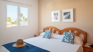 Ein ordentlich bezogenes Bett mit blauer Bettdecke, zwei Kissen mit Blättermotiv, ein Holz-Kopfteil und ein Fenster mit Blick auf Palmen und ein Gebäude.